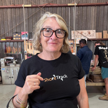 Woman wearing glasses and ShoreTrack shirt in workshop with tools and people behind